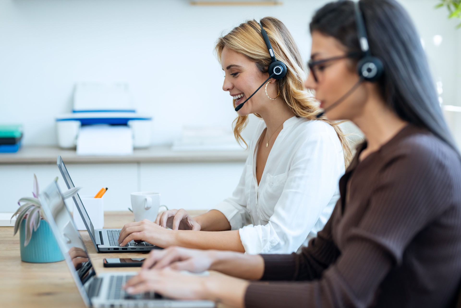 Successful attractive businesswomen working with laptops while talking with earphone on coworking place.